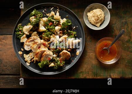 Blick von oben auf leckeres gebratenes Huhn mit Grünkohl und Blumenkohl Serviert auf Teller in der Nähe von Schüsseln mit leckeren Hummus und Chili Sauce auf rustikalem Holzbrett Stockfoto
