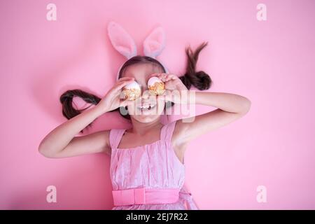 Kleines Mädchen mit Osterhasen Ohren posiert mit festlichen Ostereiern liegen auf einem rosa Studio Hintergrund. Stockfoto