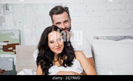 Glückliches junges Paar lächelt an der Kamera, während sie im Schlafzimmer umarmt Stockfoto