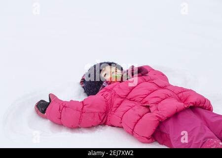 Ein kleines Mädchen in einer rosa Jacke macht einen Engel auf dem frisch gefallenen Schnee. Winter Kinder Spaß Konzept. Stockfoto