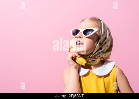 Mädchen in Kopftuch und Sonnenbrille hält Banane in der Nähe Ohr isoliert Auf Rosa Stockfoto