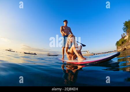 Junge glückliche Paar haben Spaß auf Stand Up Paddleboard. Aktive Paddelboarder paddeln am Meer bei Sonnenuntergang. Gesunder Lebensstil. Wassersport, SUP Surfing Tour in Stockfoto