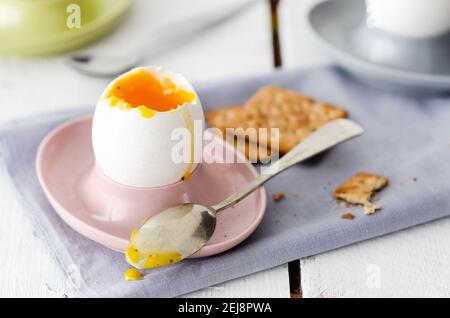 Weich gekochtes Ei in rosa, hellblau und Eierbecher mit Löffeln, einer hellblauen Serviette und Saatkräckern, im weißen Holzhintergrund. Stockfoto
