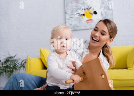 Fröhliches Kind Blick auf die Kamera, während Reiten Schaukelpferd in der Nähe Glückliche Mutter Stockfoto