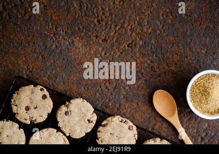 Vanille- und Schokoladenkekse auf einem schwarzen Gestell, ein Holzlöffel und Muscovado-Zucker in einer weißen Schüssel, auf einem dunklen Hintergrund. Stockfoto