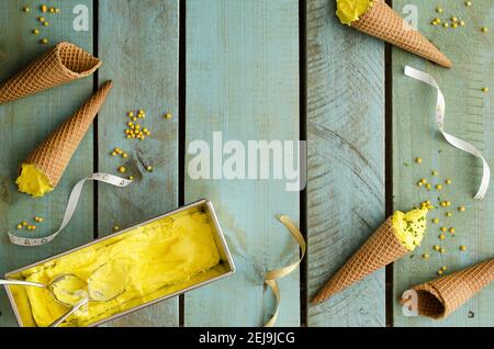 Pfirsich-Eis in einem silbernen Behälter und in Zapfen, mit gelben Bonbons und goldenen und weißen Bändern auf hellblauer Holzkulisse. Stockfoto