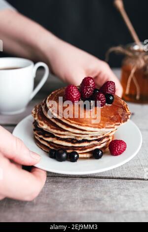 Hände halten weißen Teller mit Pfannkuchen, Beeren, Honig, Kaffeetasse auf Holztisch, Glas und Löffel. Hochwertige Fotos Stockfoto