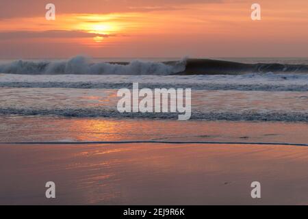 Wellen schlagen gegen Welcombe Mund Strand in Nord-Devon während des Sonnenuntergangs. Der Sonnenuntergang spiegelt sich im Sand wider. Stockfoto