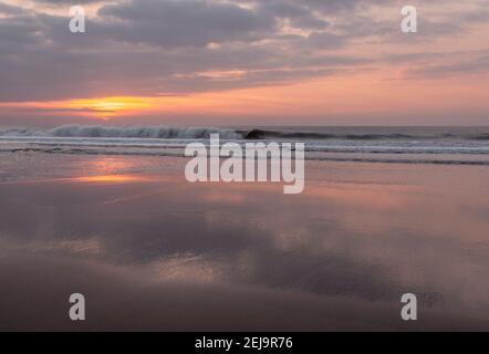 Wellen schlagen gegen Welcombe Mund Strand in Nord-Devon während des Sonnenuntergangs. Der Sonnenuntergang spiegelt sich im Sand wider. Stockfoto