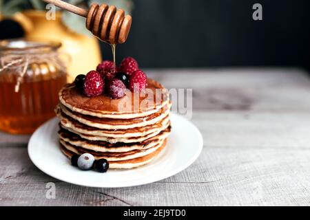 Saftige Pfannkuchen mit Beeren und Honig auf einem weißen Teller, Löffel, Glas, Holztisch, gelbe Vase mit Eukalyptus. Hochwertige Fotos Stockfoto