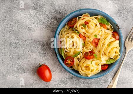 Fettuccine mit in Scheiben geschnittenen Kirschtomaten und Basilikumblättern auf einem blauen Teller auf grauem Hintergrund, daneben eine Kirschtomate. Stockfoto