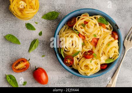 Fettuccine mit Kirschtomaten und Basilikumblättern auf einem blauen Teller auf grauem Hintergrund mit eineinhalb Tomaten, Basilikumblättern und einem Nest aus roher Pasta. Stockfoto