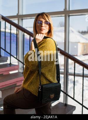 Ein stilvolles Mädchen in einem gelben Pullover, braune Hose mit einer kleinen Frauentasche posiert vor dem Hintergrund großer Fenster. Stockfoto