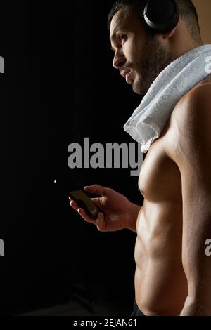 Taille nach oben Seitenansicht Porträt von bärtigen kaukasischen Männchen in Kopfhörer blicken weg und halten das Mobiltelefon in der Hand Schwarzes Zimmer Stockfoto