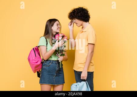 Lächelnder Teenager hält Blumen in der Nähe von Freund mit Rucksack auf gelb Hintergrund Stockfoto