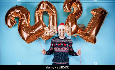 Lächelnder Mann in Pullover und rotem weihnachtsmann Hut hält golden Ballons in Form von 2021 Zahlen auf blauem Hintergrund Stockfoto