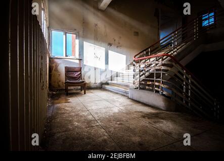 Das alte Krankenhausgebäude verlassene einen einzigen leeren Stuhl mit unheimlichen Gruselige Sonnenlichtstrahlen, die auf dunklem altem spuckten Verfallende Treppe Landung Stockfoto