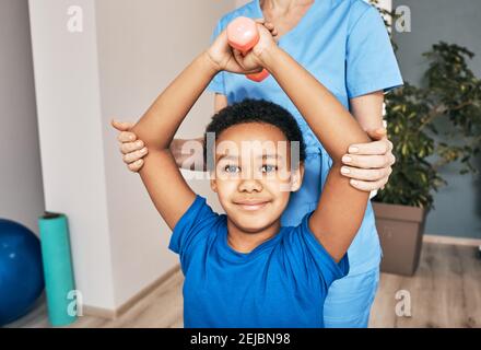 Afroamerikanischer Junge hebt Hanteln, Physiotherapeut hilft richtig zu trainieren Rehabilitation Gesundheit des Kindes Stockfoto