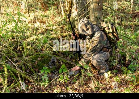 Das erschossene Birkhuhn fiel unter mit Moos bedeckte Äste, verstreute Federn lagen herum, Jäger mit Waffe hockte sich in der Nähe der Beute Stockfoto