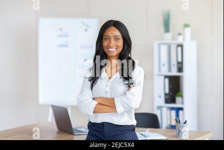 Porträt einer glücklichen afroamerikanischen Geschäftsfrau, die mit gekreuzten Armen steht Vor ihrem Schreibtisch im modernen Büro Stockfoto