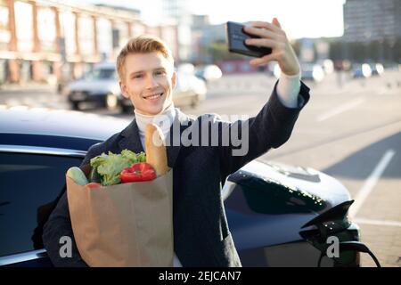 Porträt eines jungen, gutaussehenden europäischen Mannes, der Selfie-Fotos auf seinem Smartphone macht, während er sich auf sein Elektroauto stützt und den Akku bei Stadtstrom lädt Stockfoto