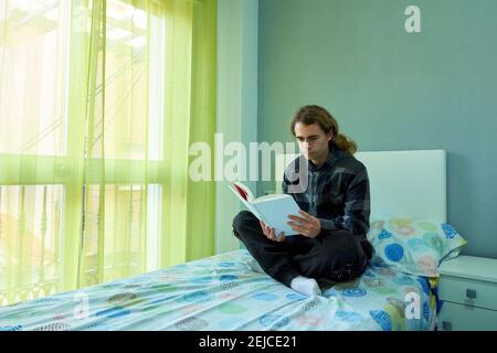 Hübscher kaukasischer Mann mit langen Haaren, die in seinem Bett sitzen Mit gekreuzten Beinen und Lesen eines Buches Stockfoto