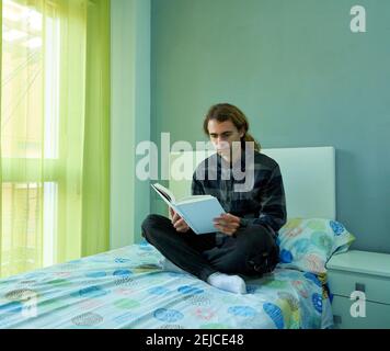 Hübscher kaukasischer Mann mit langen Haaren, die in seinem Bett sitzen Mit gekreuzten Beinen und Lesen eines Buches Stockfoto
