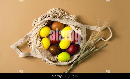 Bunte Ostereier liegen in einem Öko-Beutel auf beigem Hintergrund mit einem Bouquet aus getrockneten Kräutern, flaches Layout Stockfoto