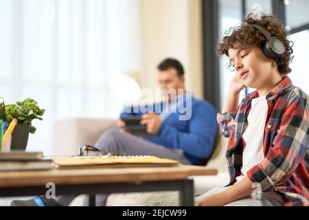 Hispanischer Schuljunge, der Kopfhörer trägt und Hausaufgaben vorbereitet, während er zu Hause am Schreibtisch sitzt. Fernstudium, Online-Bildungskonzept Stockfoto