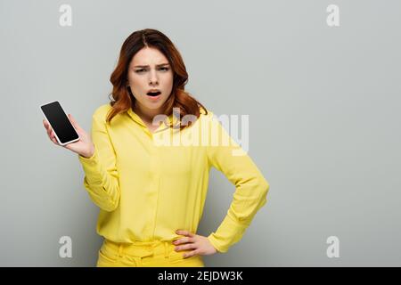Entmutigte Frau stehend mit der Hand auf der Hüfte, während das Smartphone zeigt Mit leerem Bildschirm auf grau Stockfoto