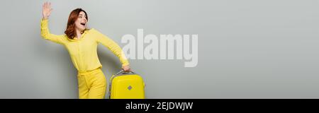 Aufgeregt Frau winkende Hand, während mit gelben Koffer auf grauem Banner stehen Stockfoto