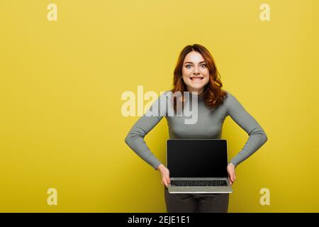 Fröhliche junge Frau mit lockigen Haaren hält Laptop mit blank Bildschirm und beißende Lippen auf gelb Stockfoto