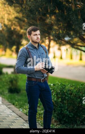 Porträt des professionellen Fotografen draußen. Mann verwenden beste Kamera und Lederband. Gutaussehender, selbstbewusster junger Mann im Hemd, der gegen die Straße steht Stockfoto