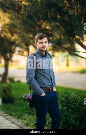 Porträt des professionellen Fotografen draußen. Mann verwenden beste Kamera und Lederband. Gutaussehender, selbstbewusster junger Mann im Hemd, der gegen die Straße steht Stockfoto