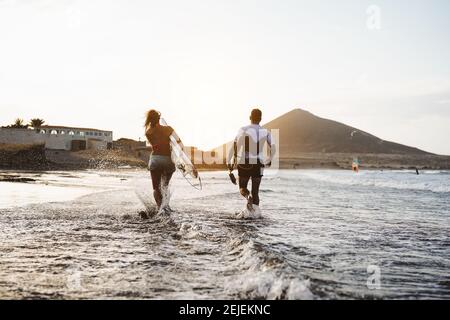 Fröhliche Surfer, die bei Sonnenuntergang im Wasser laufen - Junge Paare haben Spaß beim Surfen im Meer - Lifestyle-Konzept für Extremsport und Jugendkultur Stockfoto