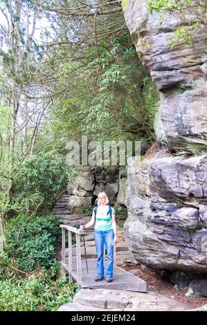 Brücke am Cragside Northumberland UK Felsen Mauern hohe Büsche Mädchen blonde Laufbahn Bahn Straße Jeans Top Stick Rucksack Garten Gärten Stockfoto