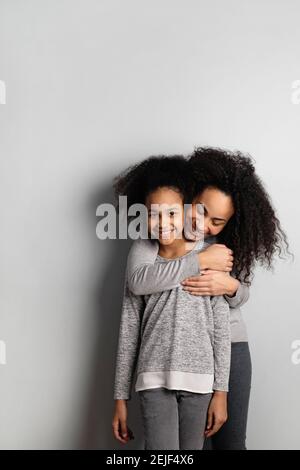 Porträt der Mutter umarmt ihre Tochter von hinten auf grauem Hintergrund mit Kopierraum. Stockfoto