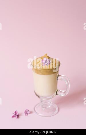 Schöner Dalgon-Kaffee-Drink. Kaffeebecher aus klarem Glas mit Schaumstoff und Fliederblüte auf einem lila rosa Hintergrund. Stockfoto