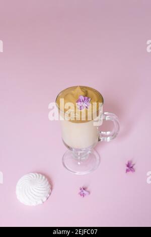 Schöner Dalgon Kaffee trinken. Kaffeebecher aus klarem Glas mit Schaumstoff und Fliederblüte auf fliederfarbenem Hintergrund. Süße zum Getränk-Marshmallow Stockfoto