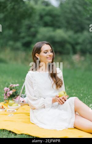 Lächelndes kaukasisches Teenager-Mädchen im Freien sitzen. Kreative künstlerische junge Frau trägt Freizeitkleidung mit einem Picknick mit Apfel am Sommertag. Stockfoto