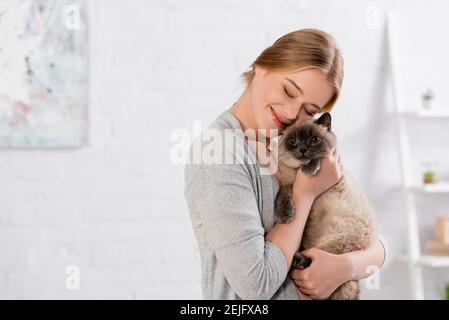 Lächelnde Frau mit geschlossenen Augen umarmt siam Katze Stockfoto