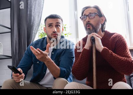 Junger hispanischer Mann zeigt mit der Hand, während er Fernsehen in der Nähe Erstaunt Vater Stockfoto