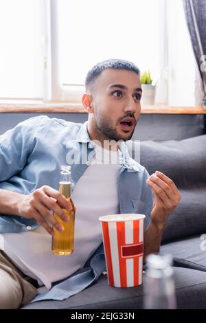 Staunender hispanischer Mann mit einer Flasche Bier beim Fernsehen und beim Essen von Popcorn zu Hause, verschwommener Vordergrund Stockfoto