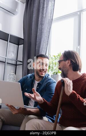 Bärtiger Mann mit Gehstock, der auf Laptop zeigt, nahe aufgeregt hispanischer Sohn Stockfoto