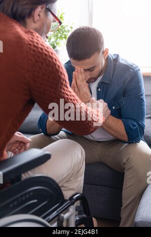 Behinderter Mann, der die Hand des deprimierten hispanischen Sohnes berührt, der mit sitzt Gebeugt Stockfoto