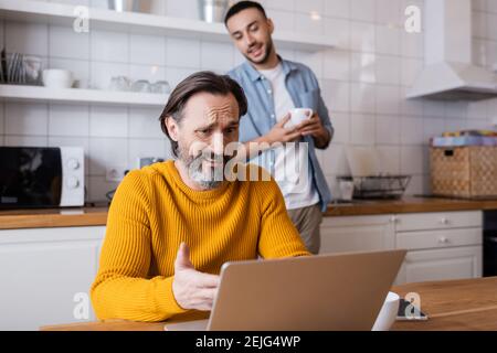 Skeptischer Mann, der auf einen Laptop zeigt, mit dem der hispanische Sohn fast lächelte tasse auf unscharfem Hintergrund Stockfoto