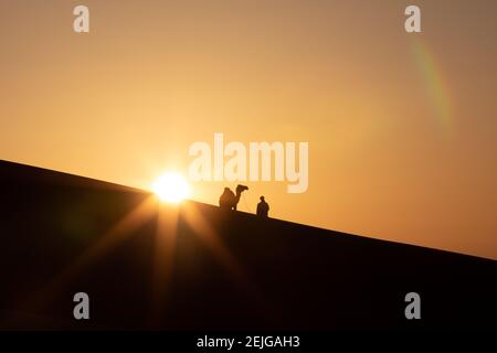 Silhouette eines Nomadenmannes mit seinem Kamel in Sanddünen bei Sonnenuntergang. Wüste Liwa, Abu Dhabi, VAE. Stockfoto