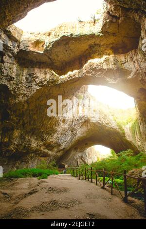 Devetashka Höhle in Bulgarien - natürliche Attraktion. Hohe Bögen einer riesigen Steinhöhle mit runden Löchern an der Spitze, eine Touristenstraße mit einem Zaun in der Stockfoto