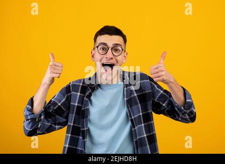 Funky junge Kerl in Brille zeigt Daumen nach oben mit beiden Händen, Genehmigung von etwas auf orangen Studio Hintergrund Stockfoto