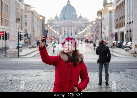 Mädchen in rotem Kleid und roter Baskenmütze nimmt ein Selfie mit dem Hintergrund des Petersdoms im Vatikan. Stockfoto